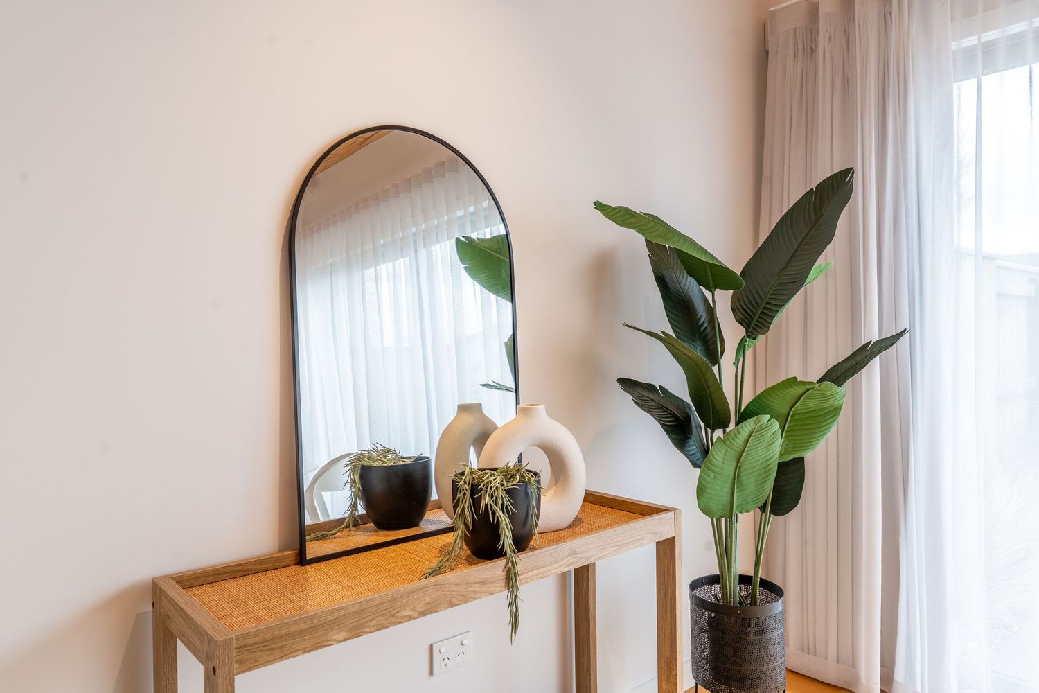 Wooden console table with arched mirror, white vases, and large indoor plant by window.