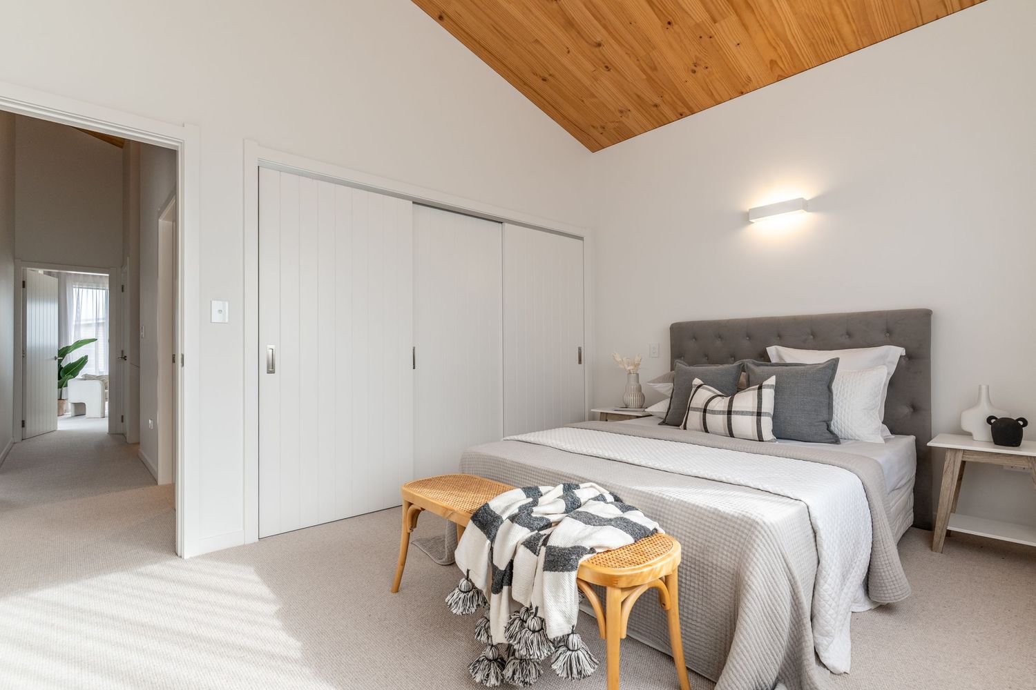 Bright bedroom with timber vaulted ceiling, grey upholstered headboard, and sliding wardrobe.