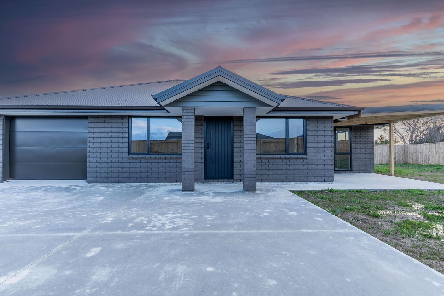 Modern brick home with double garage, gabled roofline, and dramatic sunset sky backdrop.