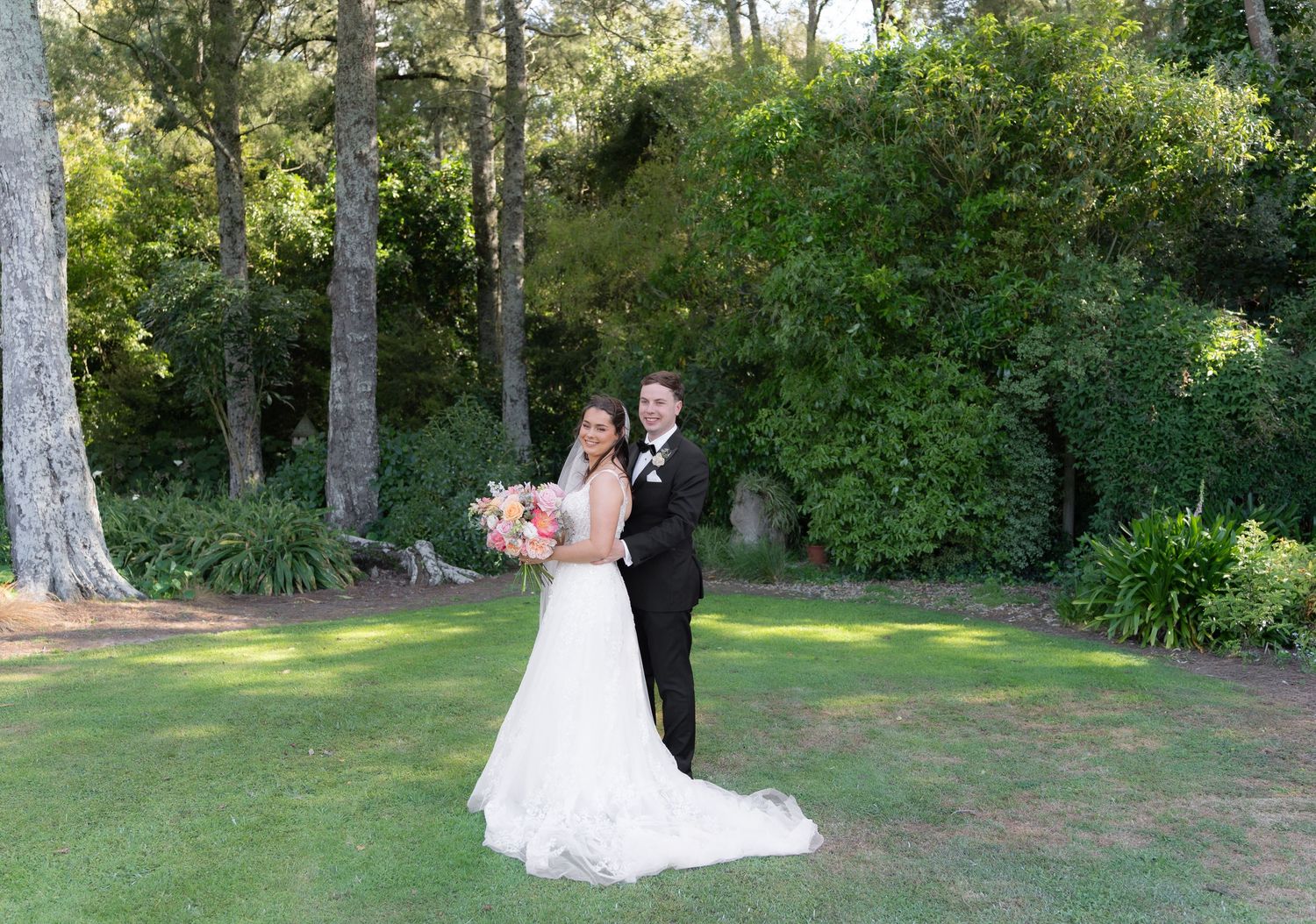 Bride and groom posing together on lush green lawn surrounded by tall trees and garden greenery on their wedding day.