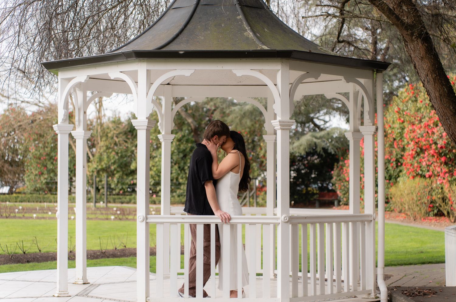 Couple sharing a romantic kiss inside a classic white wooden gazebo surrounded by autumn gardens.