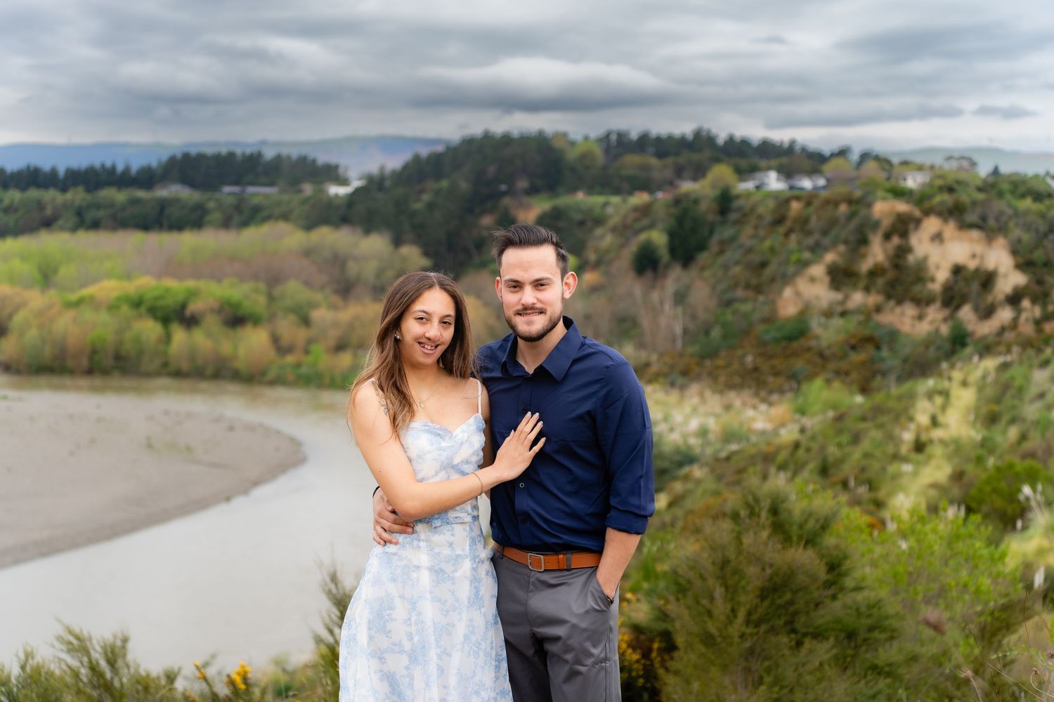 Smiling couple posing together on a hilltop overlooking a winding river under a cloudy sky.