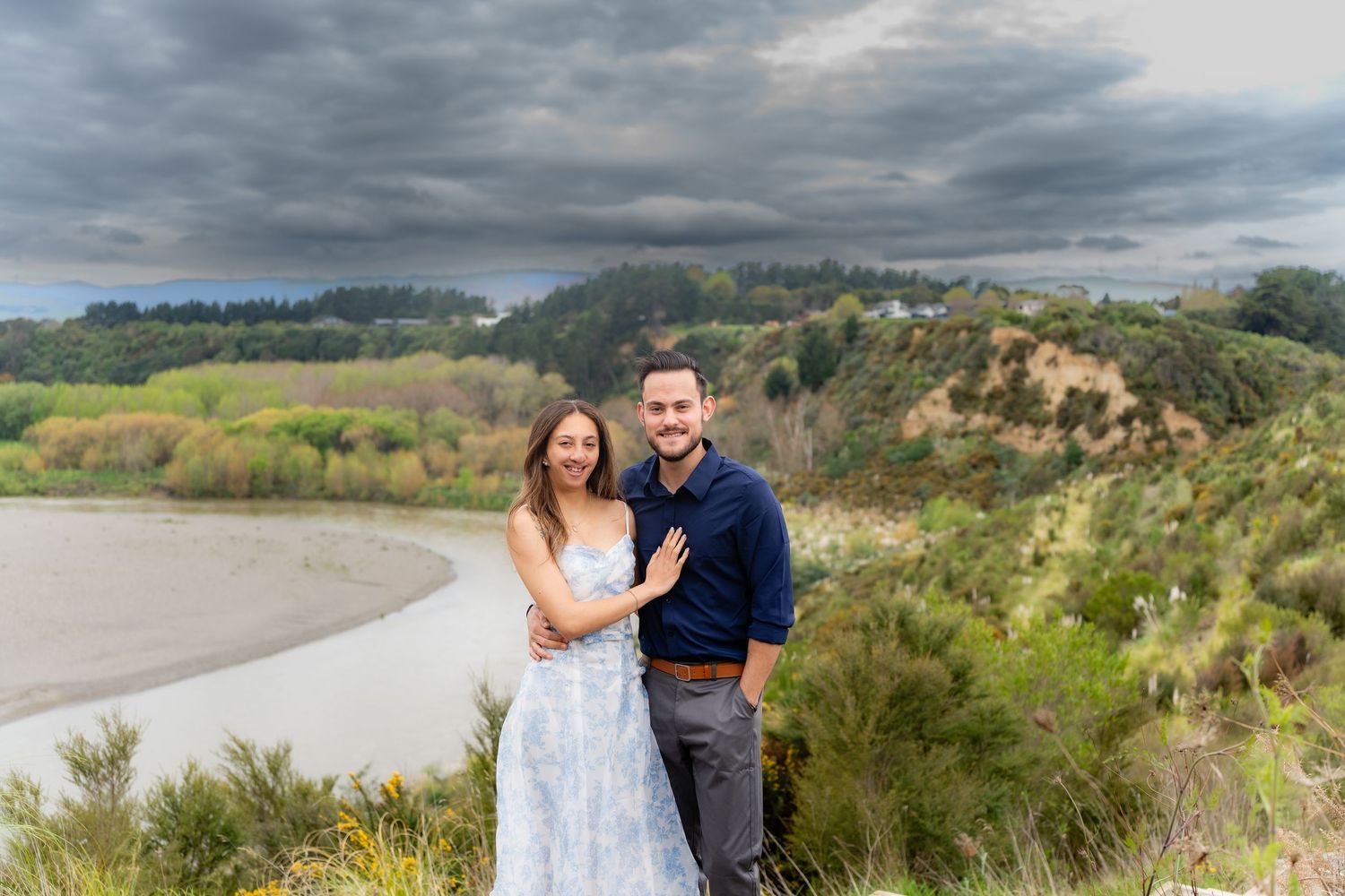 Engaged couple posing on scenic overlook with river valley and stormy sky behind them.