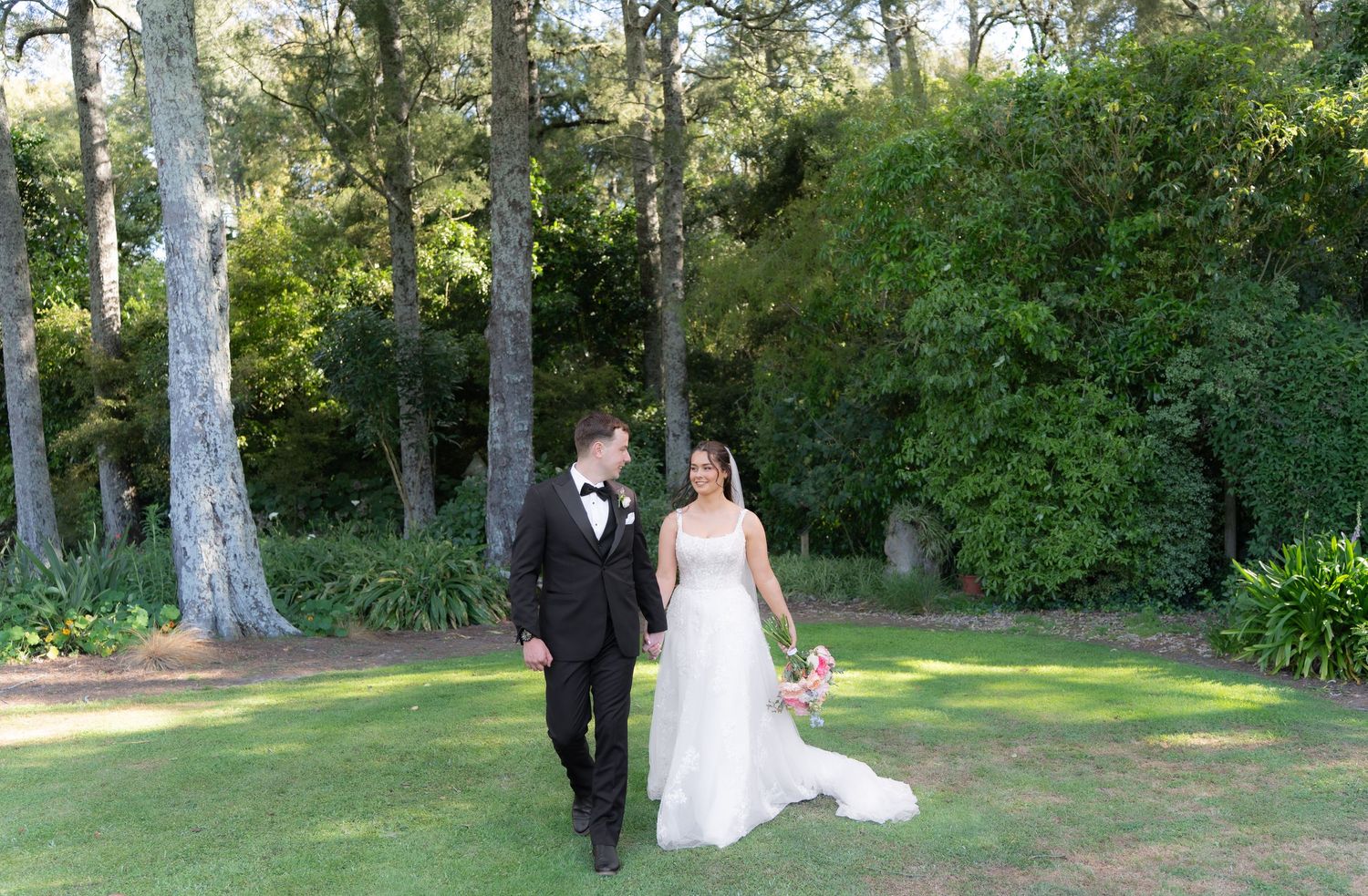 Newlywed couple strolling through scenic garden grounds with tall pine trees and lush greenery on their wedding day.