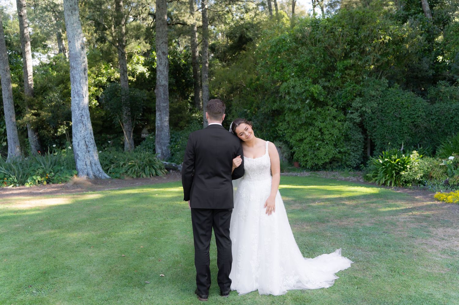Bride in white gown leaning on groom in black tuxedo during an intimate first look moment in a garden setting.