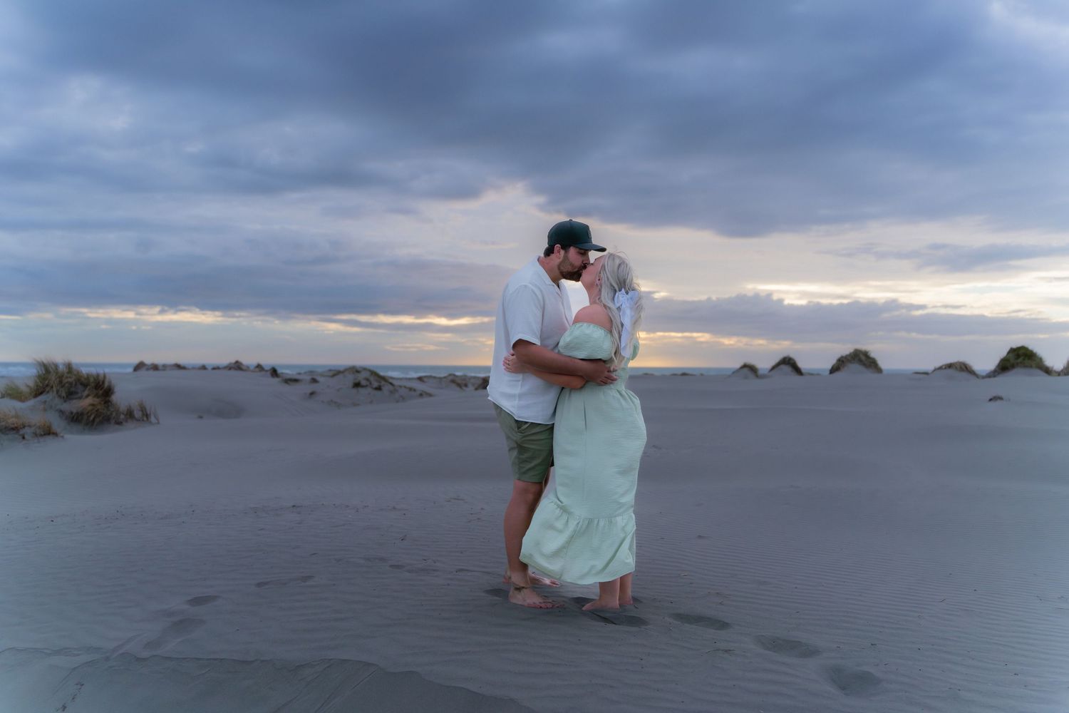 Couple embracing romantically on sand dunes at dusk under a dramatic cloudy sky.