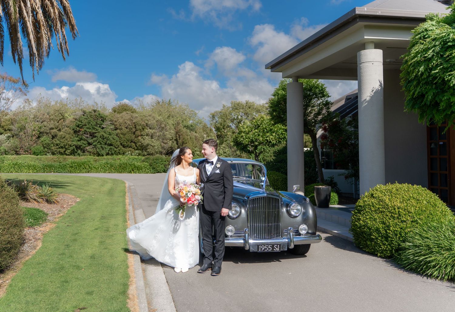 Bride and groom standing together beside a vintage car outside a grand wedding venue.