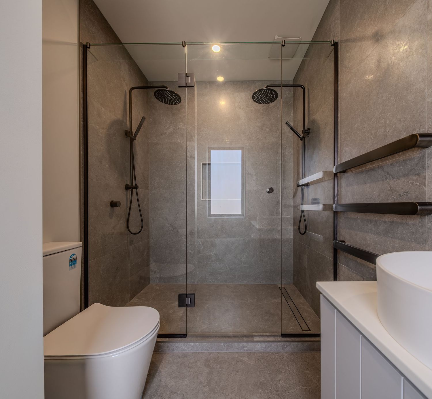 Modern bathroom with glass shower enclosure, dual rain heads, gray marble tiles, toilet, and white vanity.