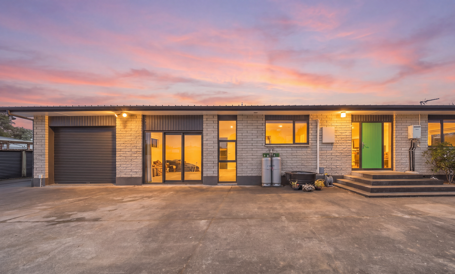 Modern brick home exterior at sunset with glowing interior lights, green front door, garage, and vibrant pink sky.