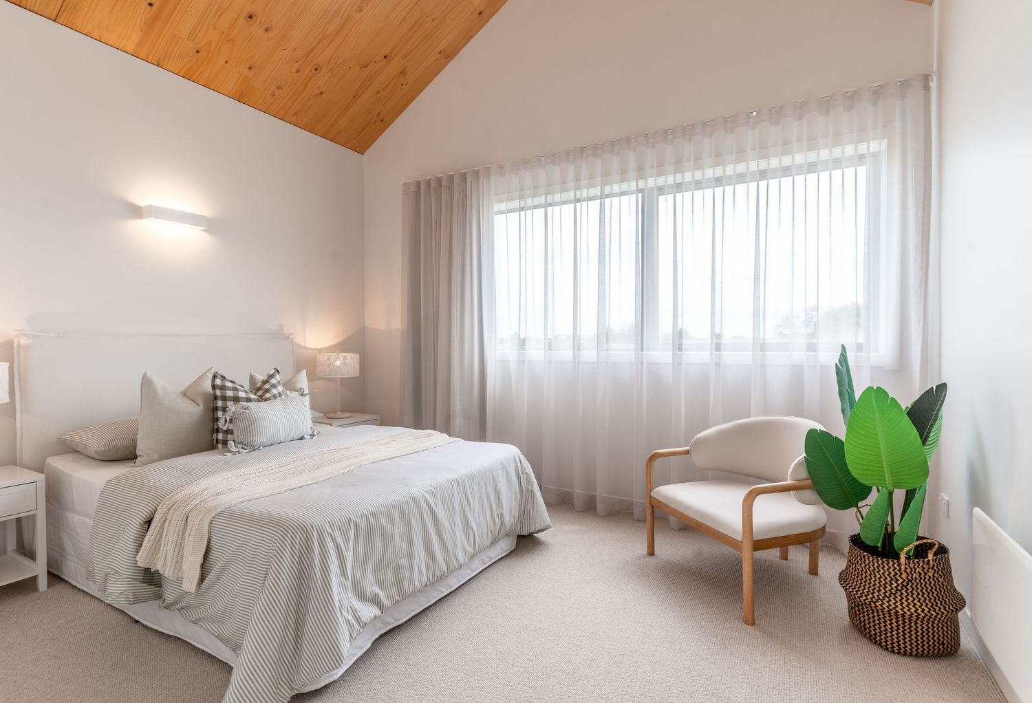 Bright white bedroom with vaulted wood ceiling, sheer curtains, neutral bedding, and a rattan chair beside a potted plant.