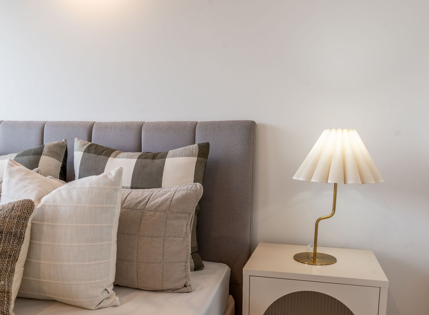Close-up of grey upholstered bed with layered neutral cushions and gold bedside lamp.