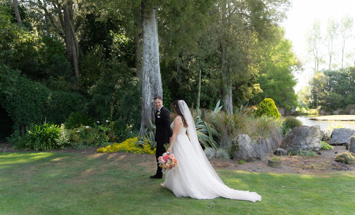 Bride in flowing white gown and groom in black suit posing near tall trees and colorful garden landscaping.