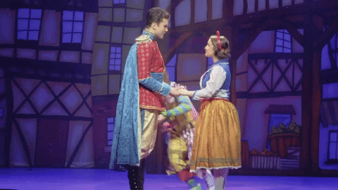 Two performers in royal costumes hold hands on a colorful theatrical stage set during a fairy tale ballet production.