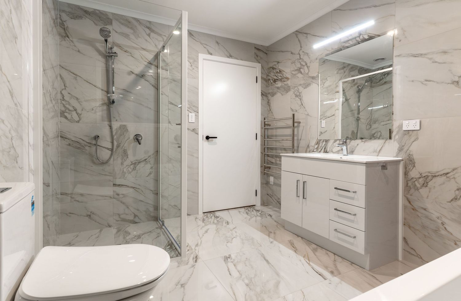 Luxurious marble-tiled bathroom with glass shower, white vanity, and heated towel rail.