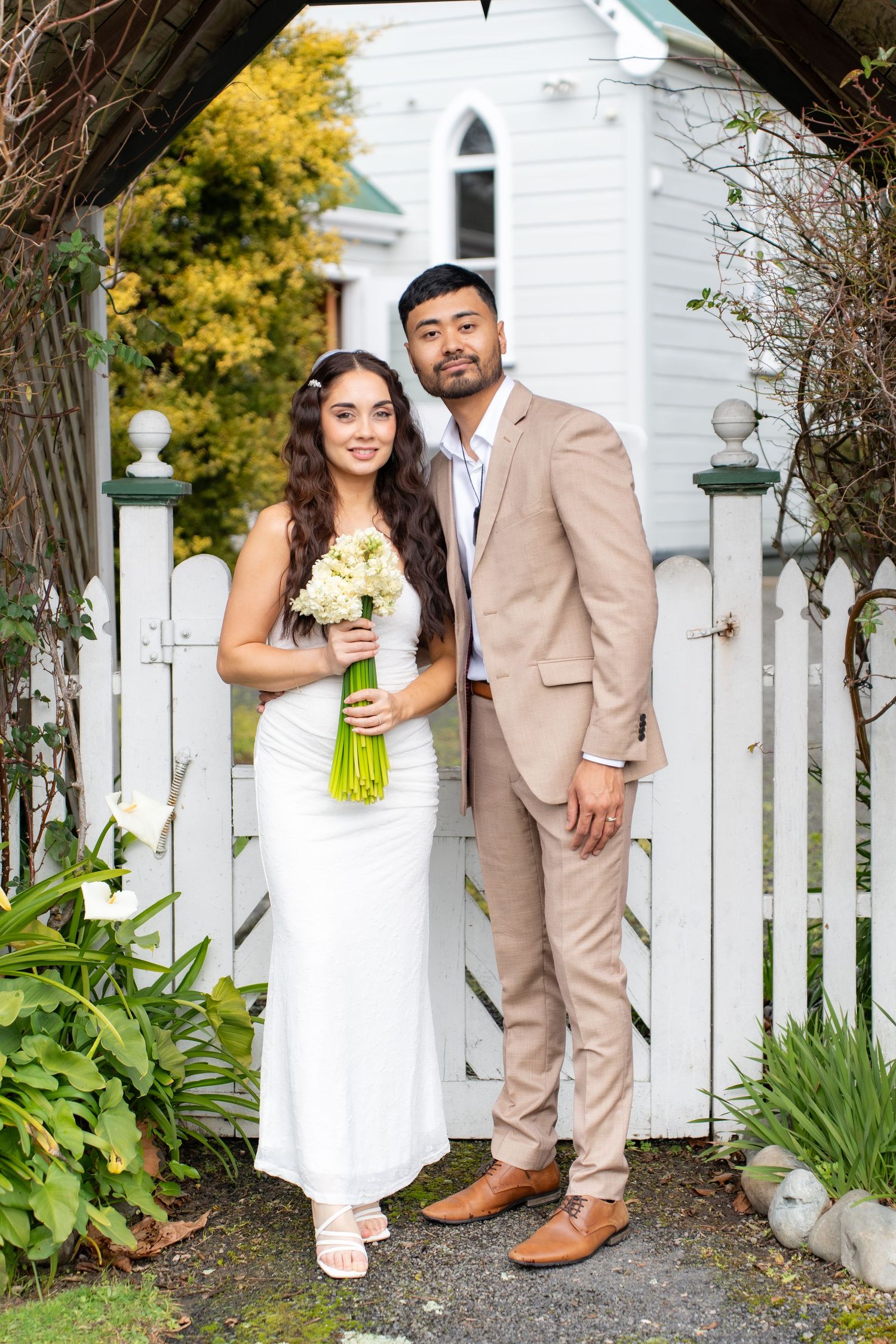 Bride and groom posing at white picket gate of historic chapel with bridal bouquet.