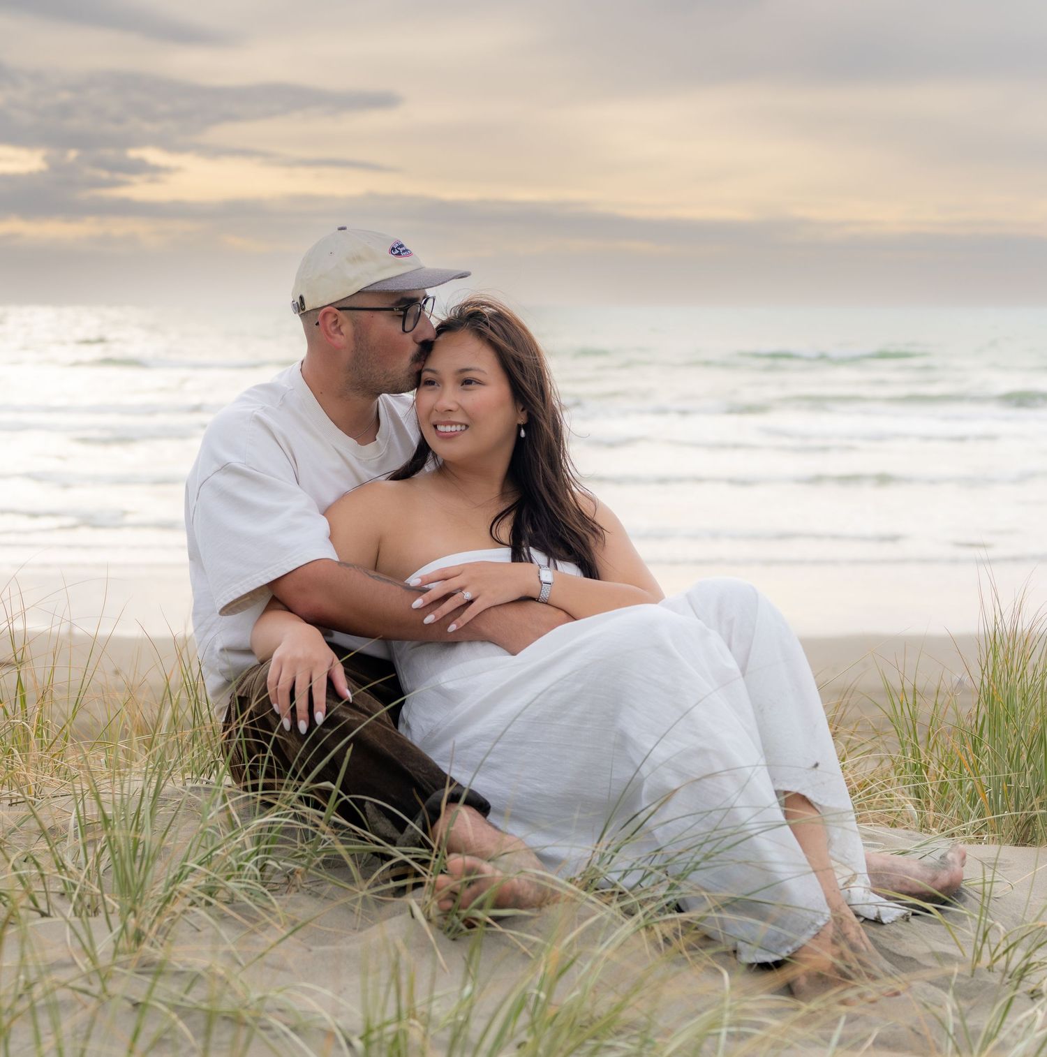 Couple sitting together on beach sand at sunset with ocean waves in background.
