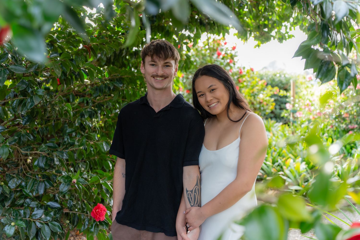 Smiling couple holding hands while standing beneath lush green foliage and red flowering plants.