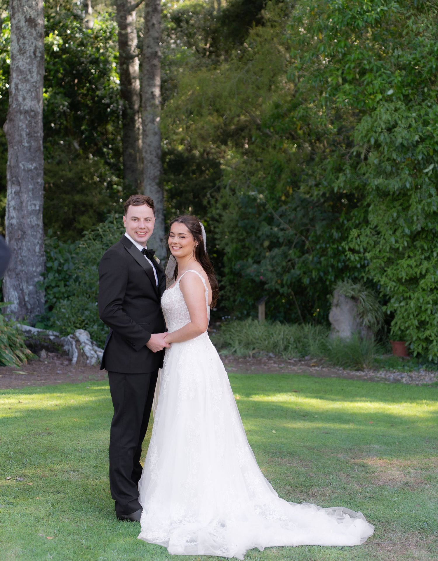 Bride and groom holding hands and smiling on green lawn with tall trees behind them.