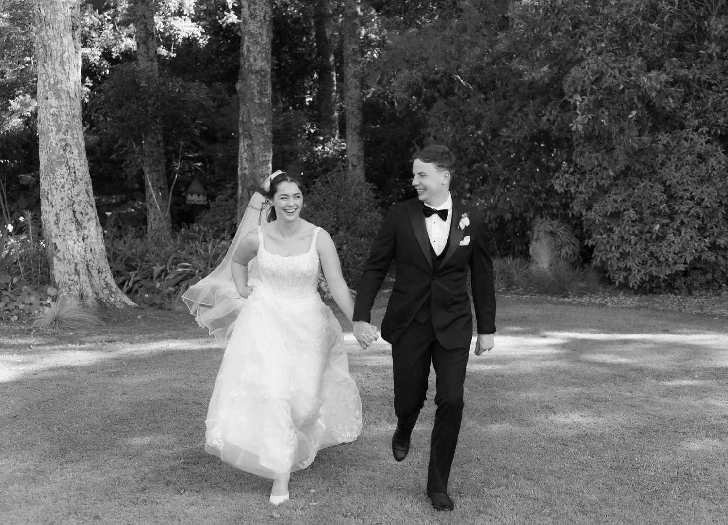 Black and white photo of happy bride and groom holding hands and walking through a tree-lined garden path.