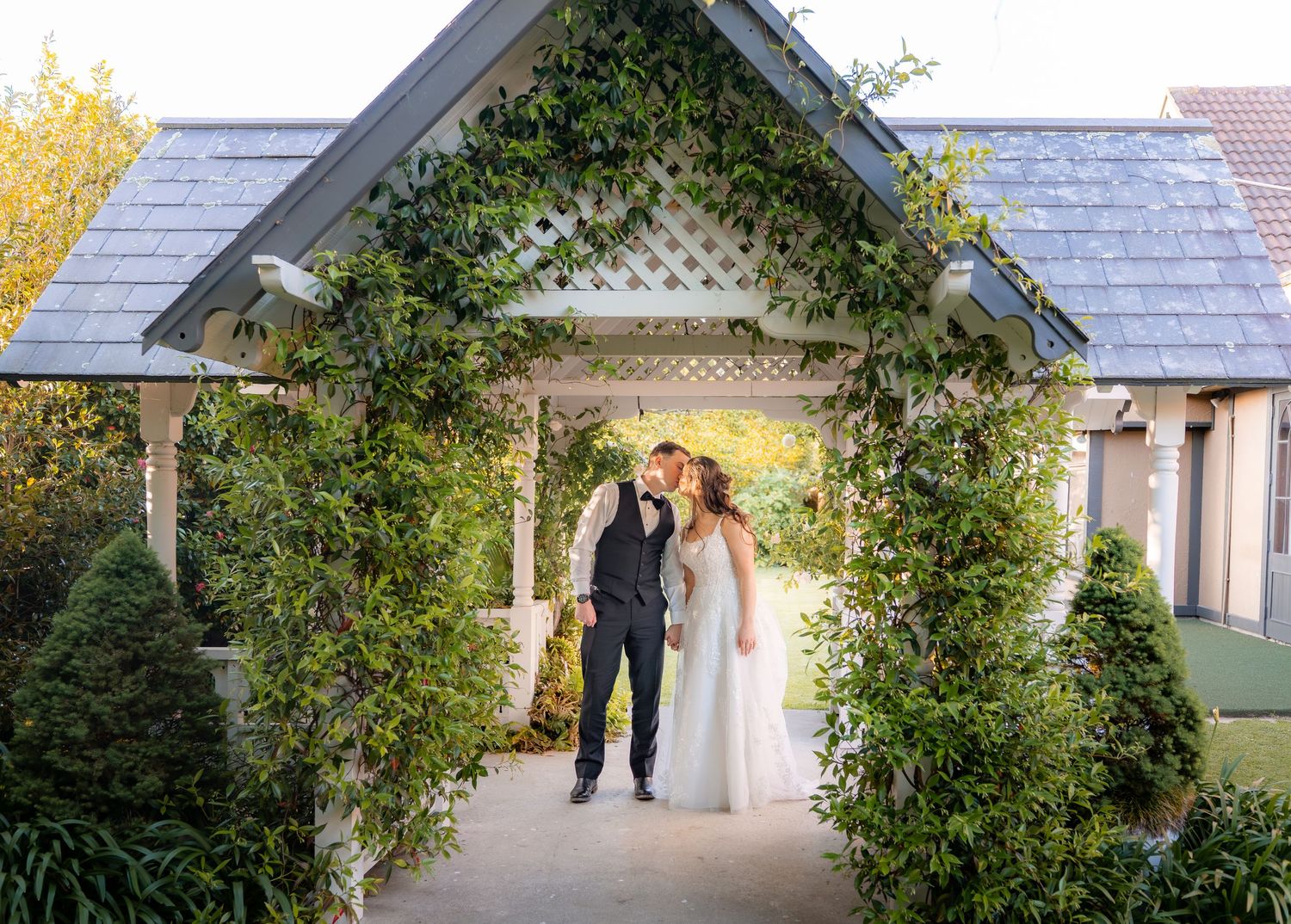 Wedding couple sharing a romantic moment under an ivy-covered white lattice pergola entrance at an elegant garden venue.