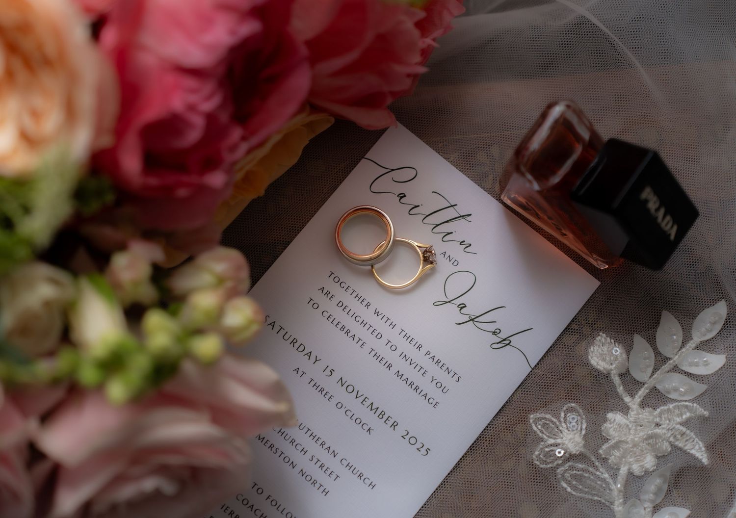 Wedding flat lay featuring gold rings, invitation, pink florals and perfume bottle.
