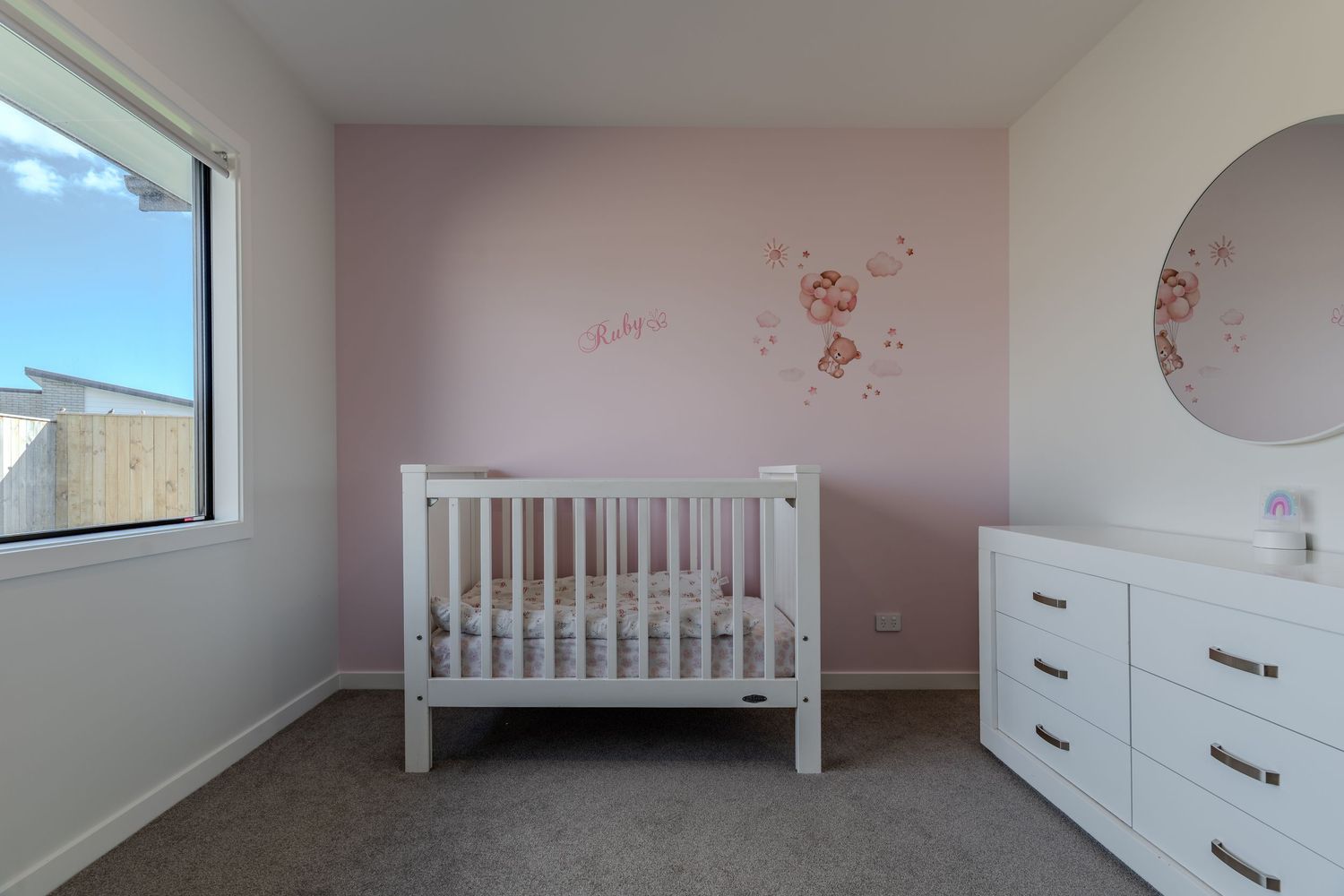Pink nursery with white crib, dresser, round mirror, and floral wall decals on a soft pink accent wall.