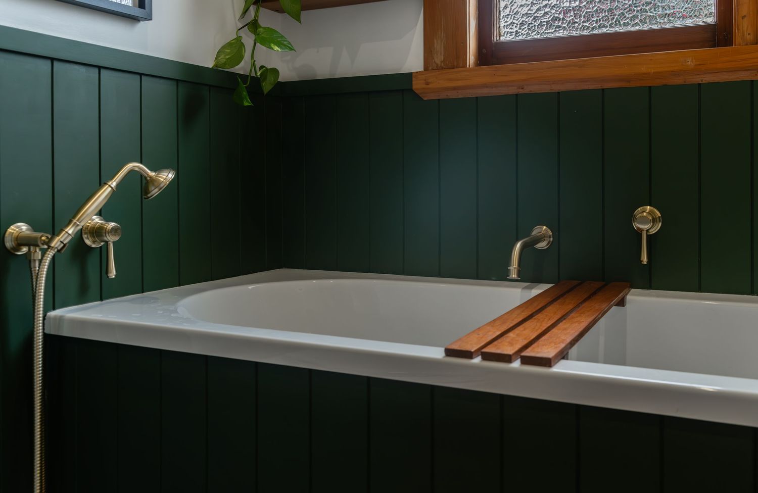 Dark green paneled bathroom with a white soaking tub, wooden bath caddy, and brass fixtures beside a frosted window.