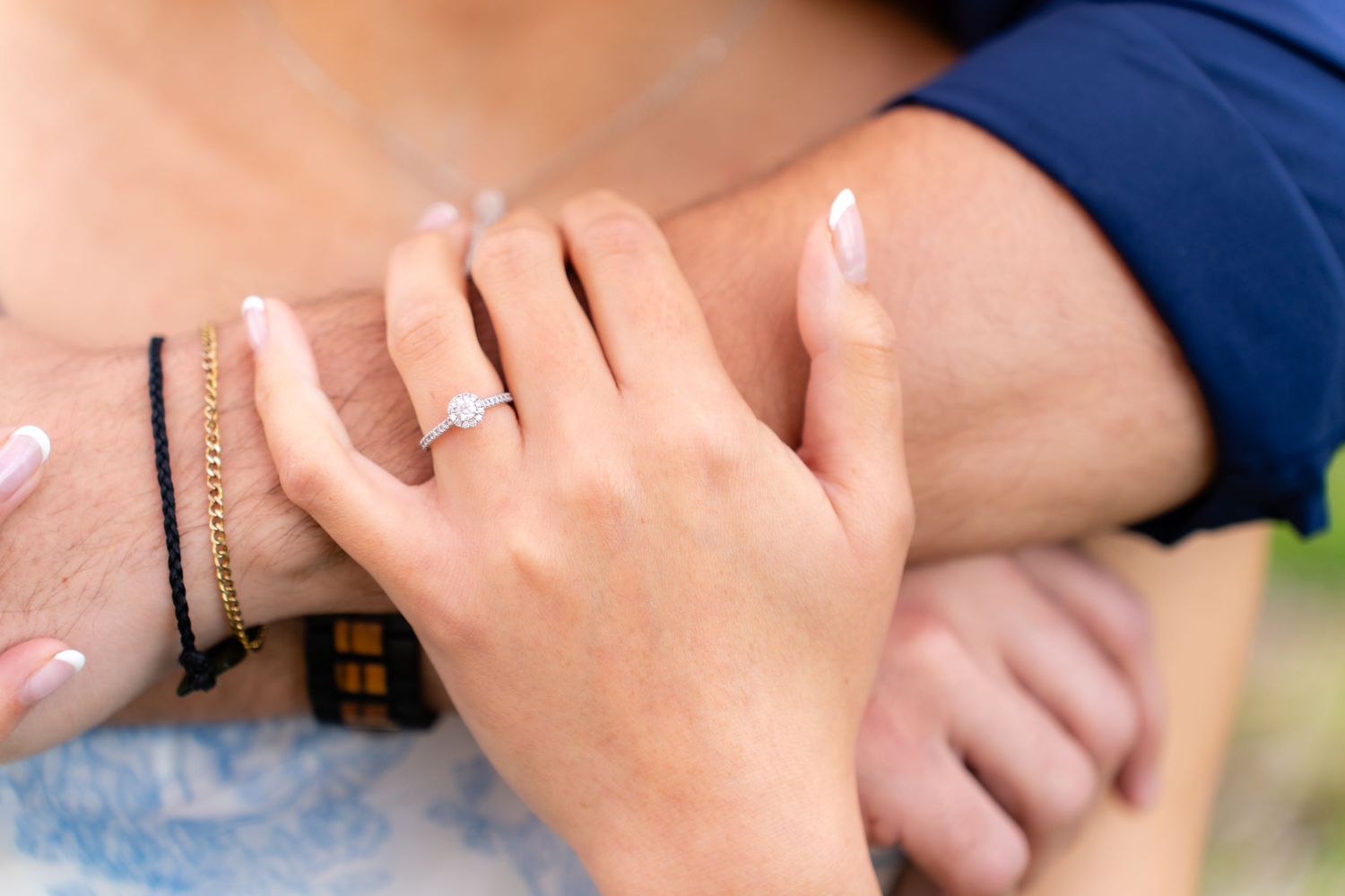 Close-up of womans hand with solitaire diamond ring resting on mans arm outdoors.