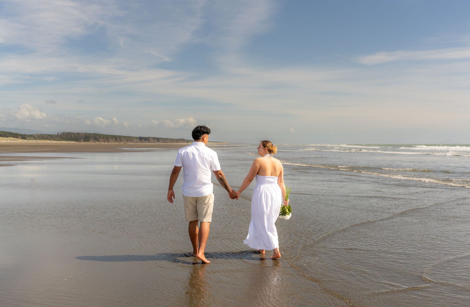 Couple holding hands walking along a sunny beach with blue skies and gentle waves.