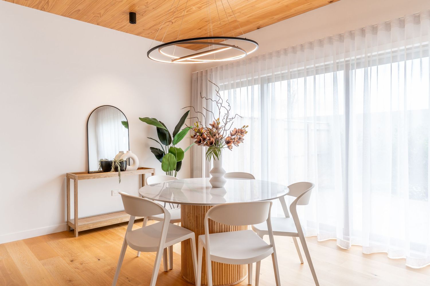 Modern dining room with round white table, wood chairs, oval mirror, and sheer curtains in a bright airy space.
