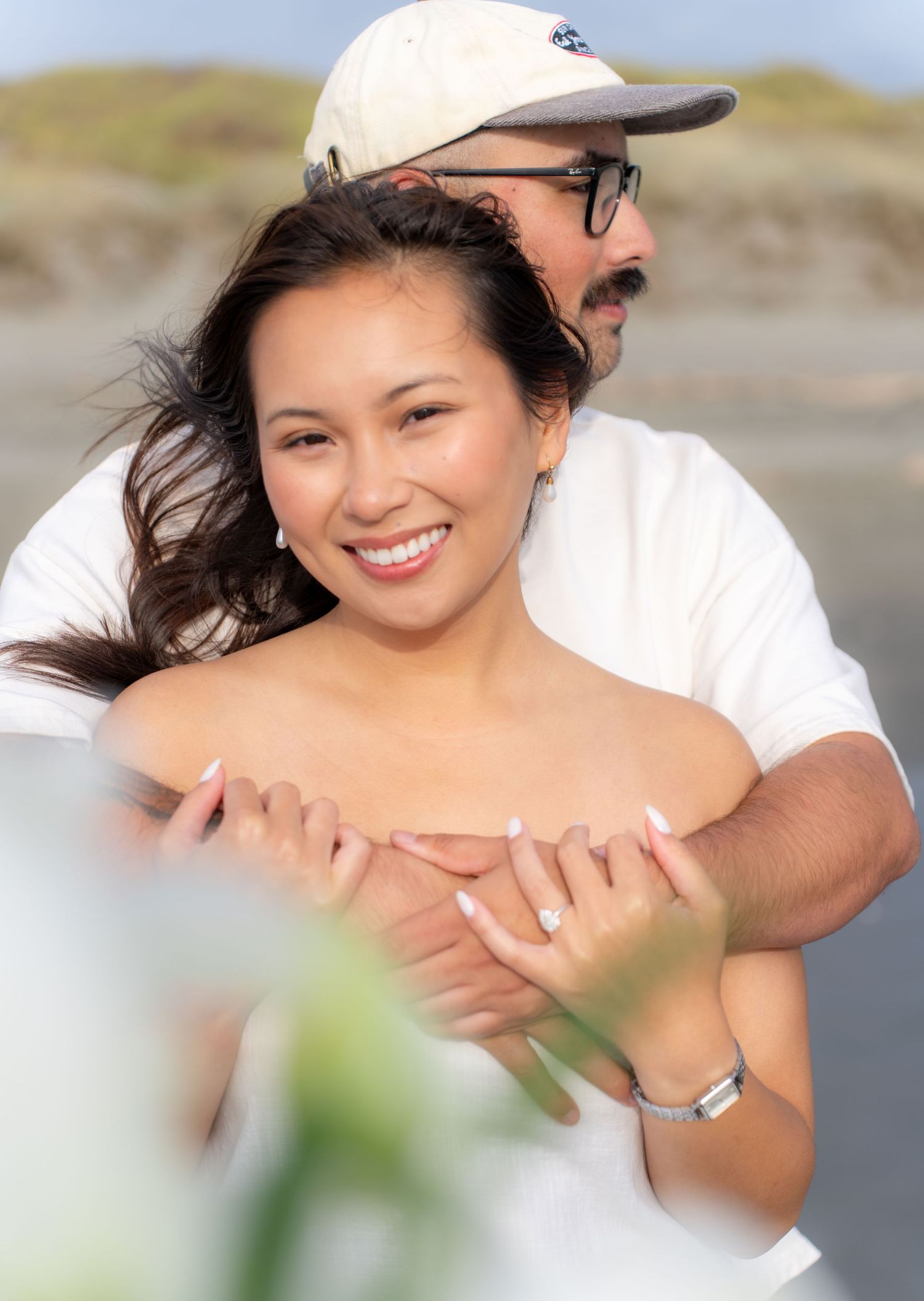 Smiling woman showing engagement ring while her partner embraces her from behind at a beach.