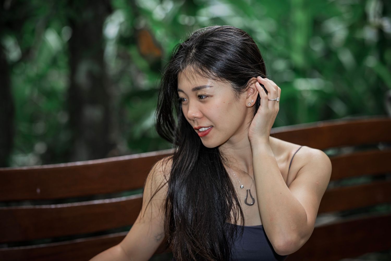 Person with long dark hair wearing black top sits on wooden bench touching hair while smiling outdoors.