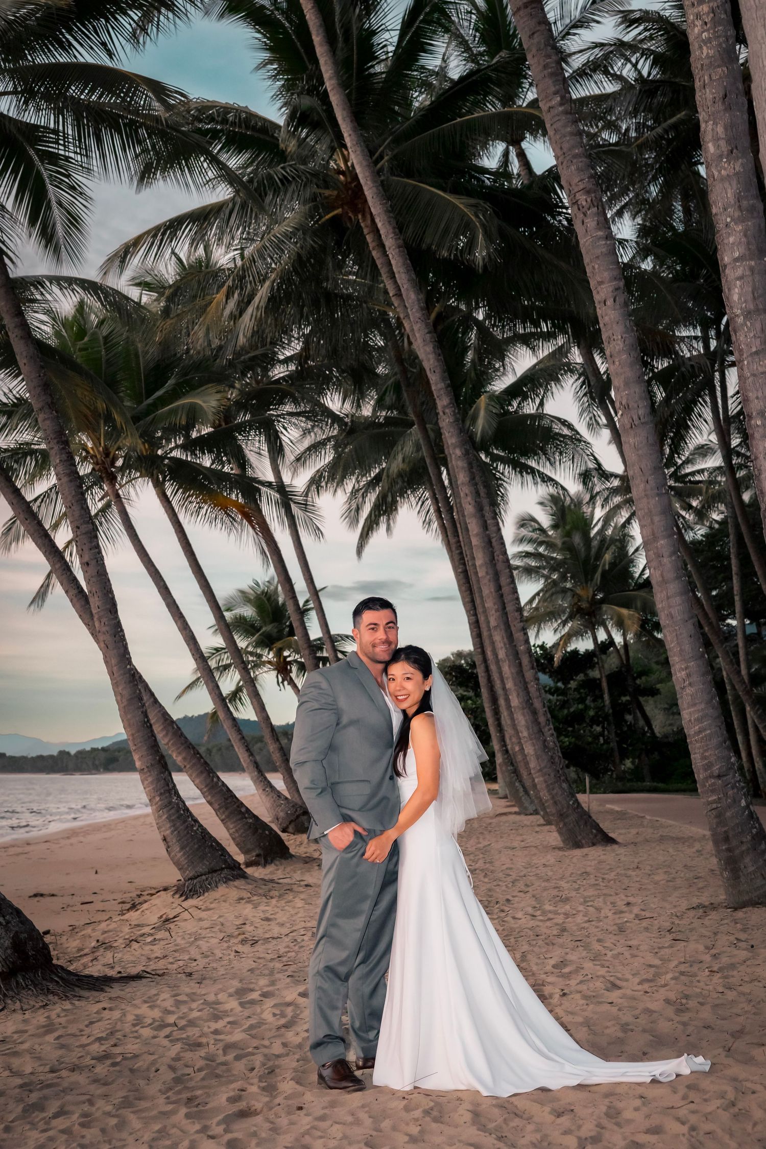 A romantic beach wedding couple poses among palm trees at sunset on a tropical shoreline.