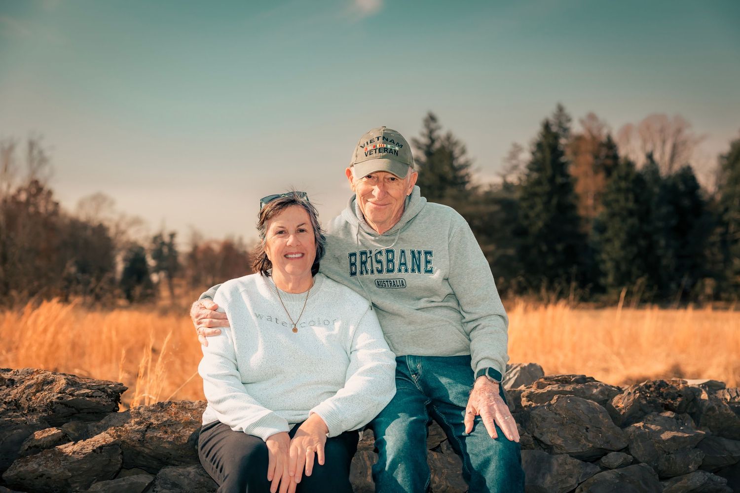 A couple sits by the water at sunset wearing blue and white casual attire.