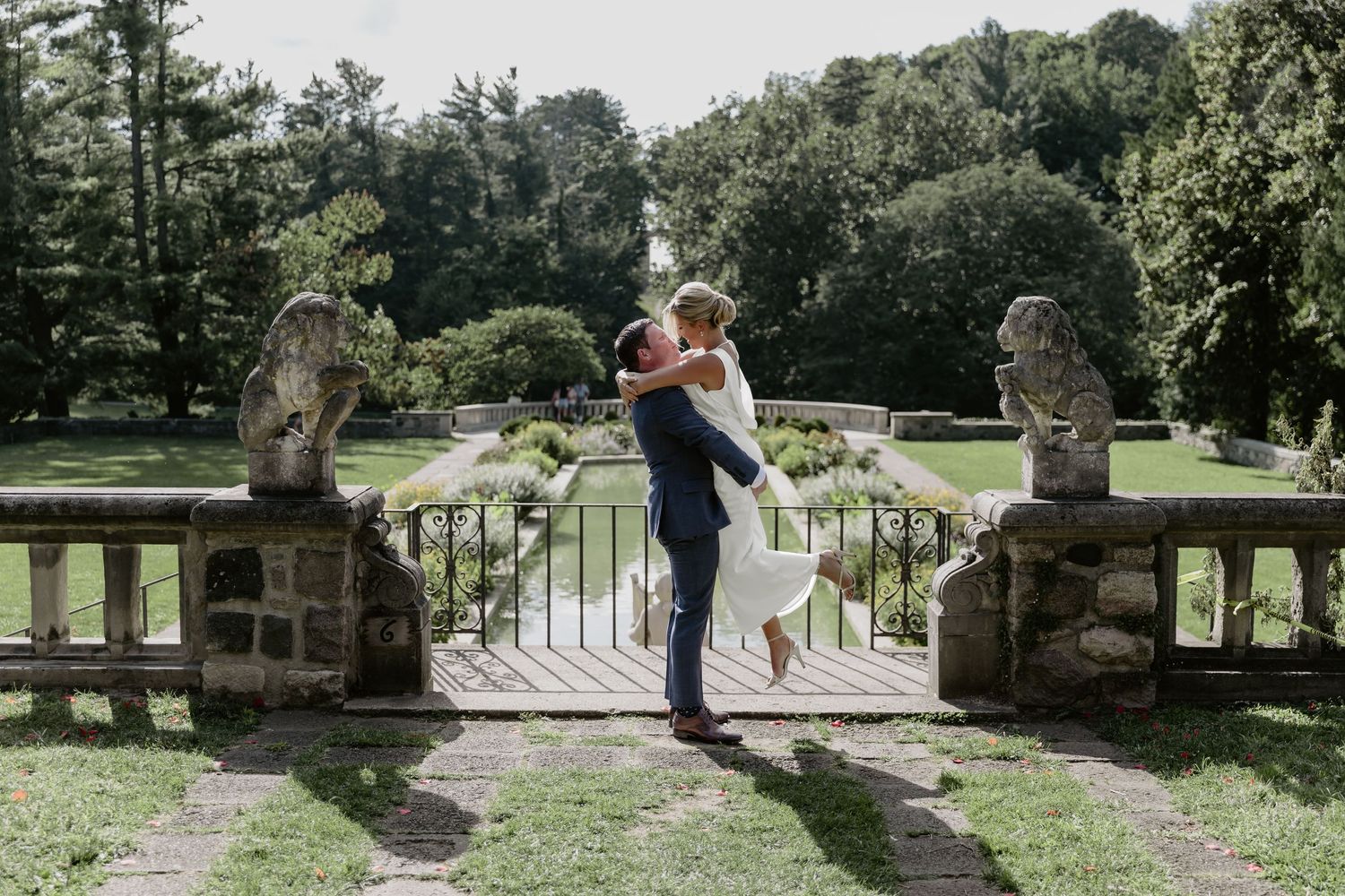 A romantic embrace on a stone bridge surrounded by lush gardens and antique statues.
