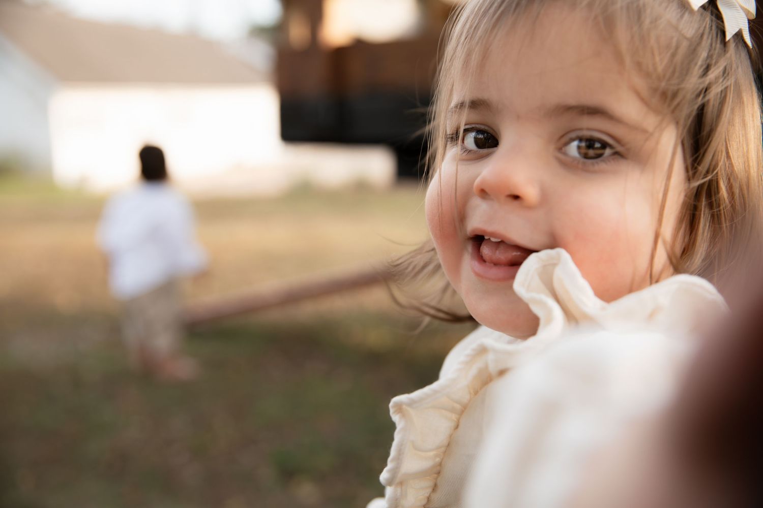 A small child in a white dress smiles outdoors at sunset while someone walks in the background.