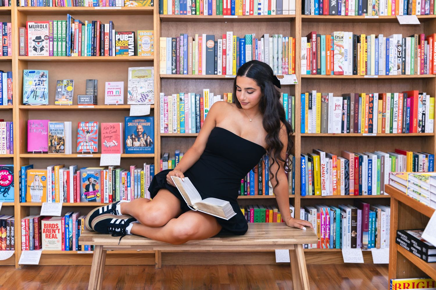 Person in black dress relaxes on wooden bench surrounded by library bookshelves.