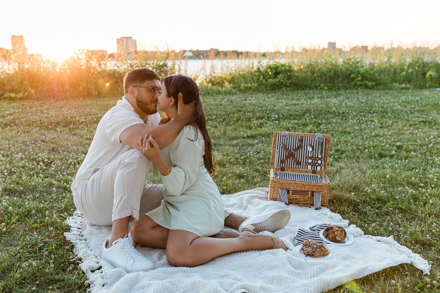 A romantic sunset picnic scene with a couple sitting on a white blanket with a picnic basket and snacks.