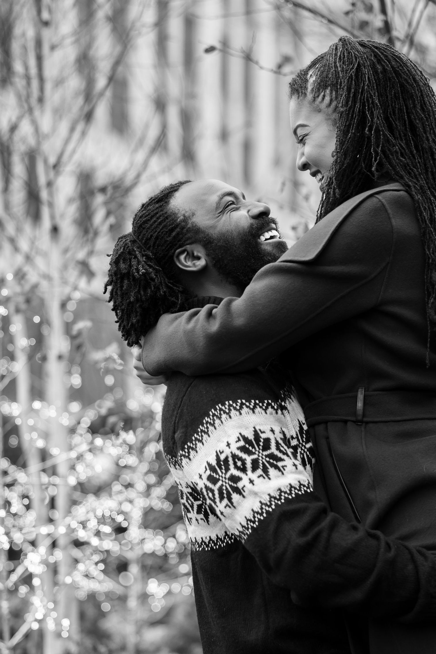 Couple shares a joyful embrace outdoors during winter with twinkling lights in the background.