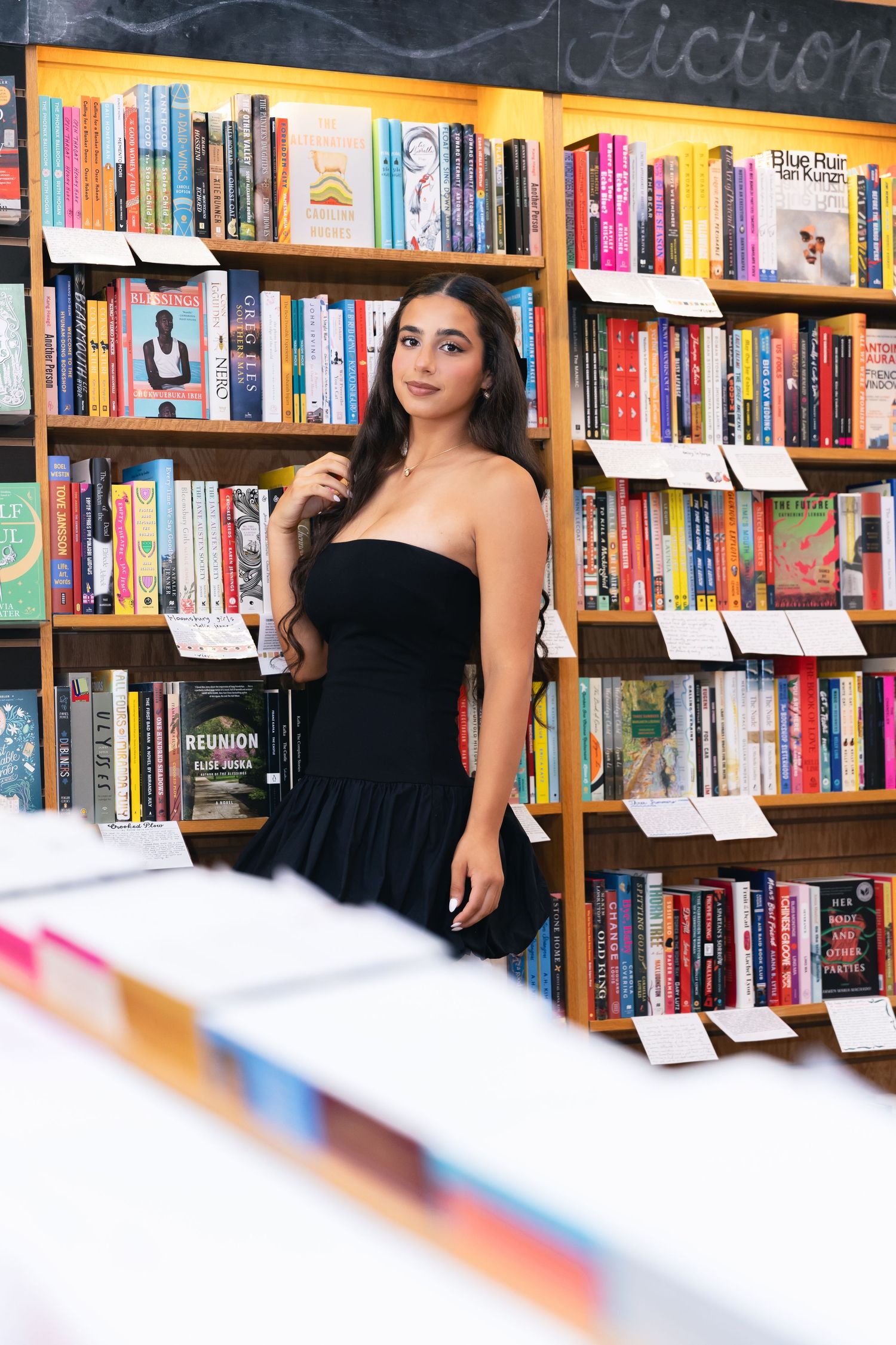 Woman in black dress standing between rows of bookshelves in library.