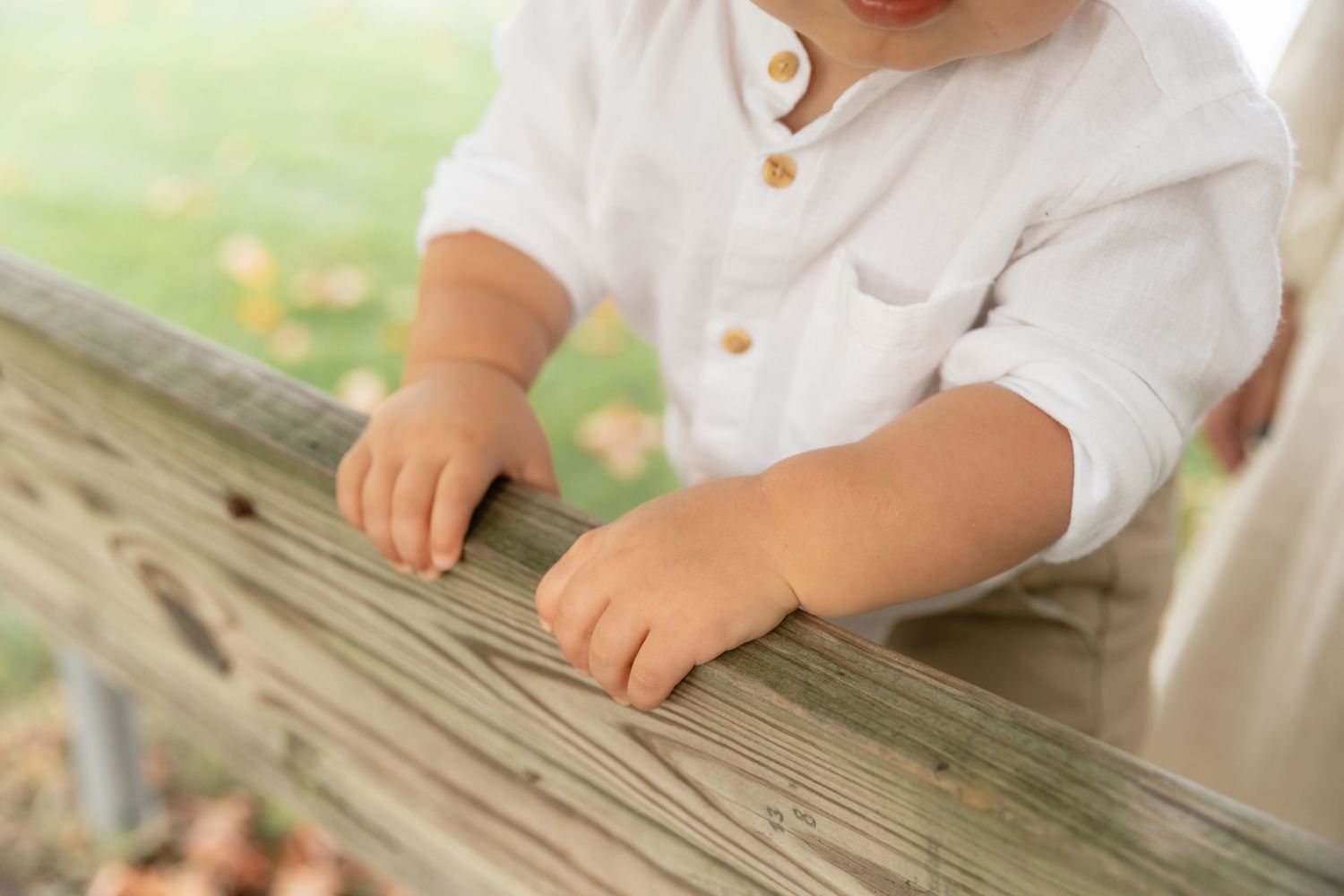 Small hands in a white button-up shirt rest on weathered wooden railing outdoors on a sunny day.