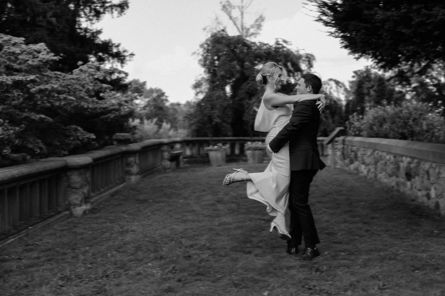 A black and white romantic lift pose on a stone bridge in a garden setting.