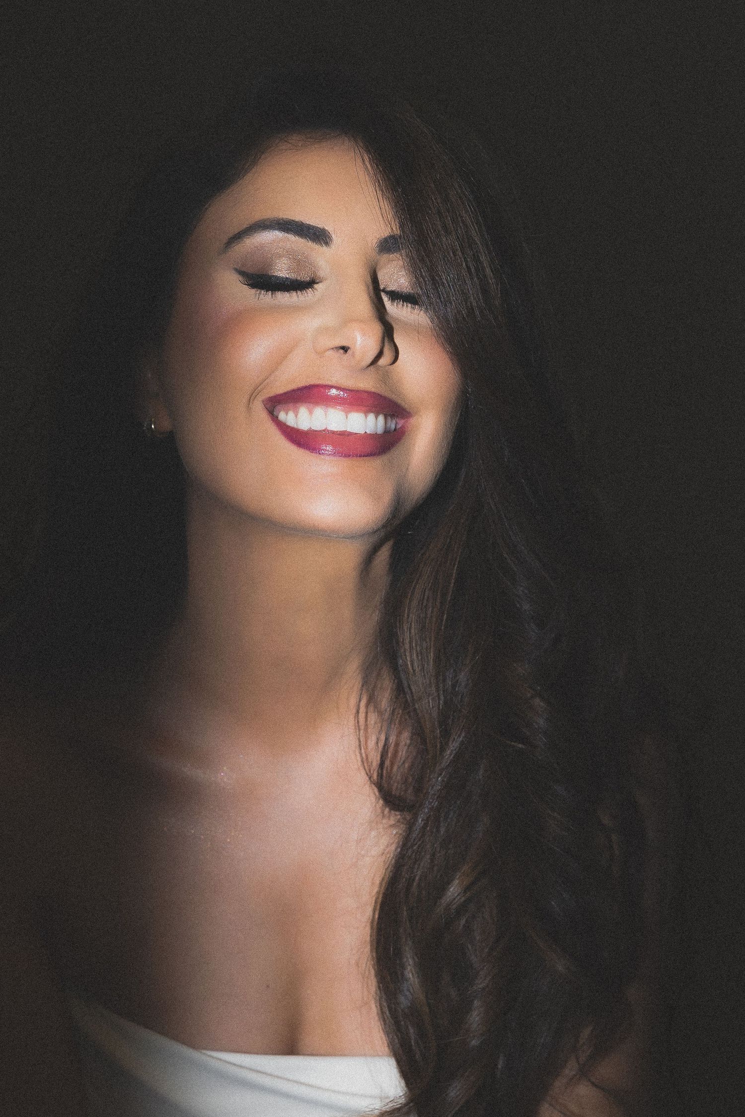 Portrait in dramatic lighting showing a natural smile, long dark hair, red lipstick and glowing makeup against black background.