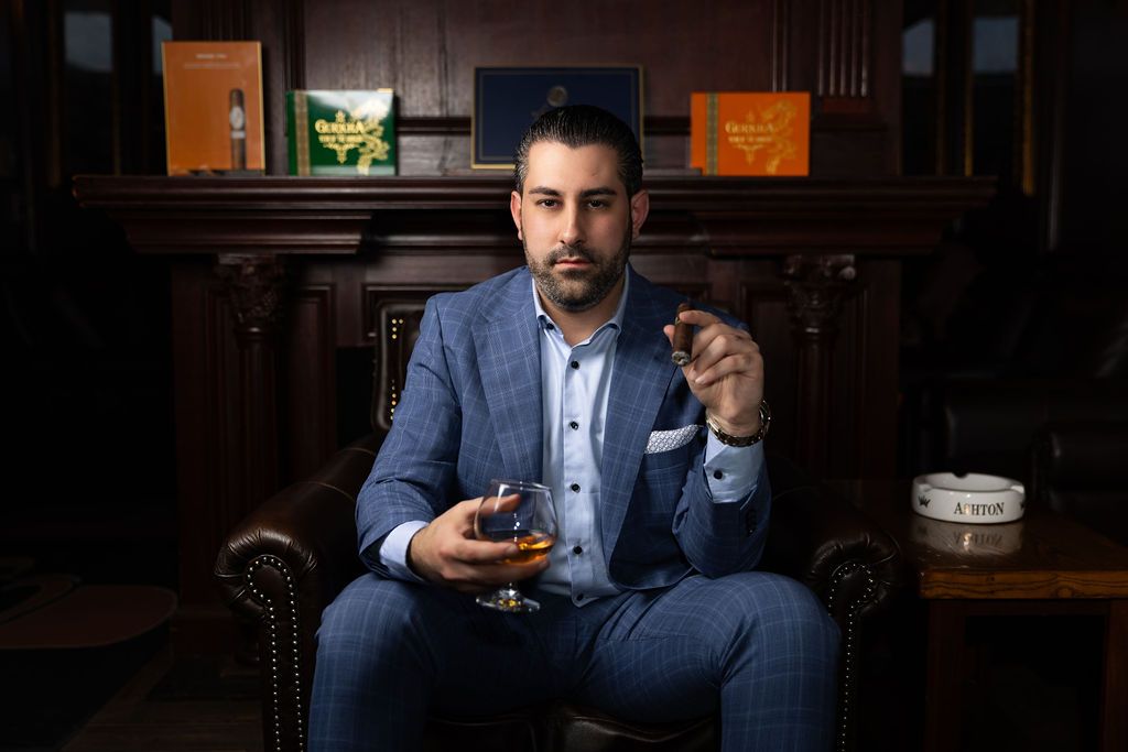 A suited person sits in a leather armchair holding a glass of whiskey and phone in a dimly lit lounge setting.