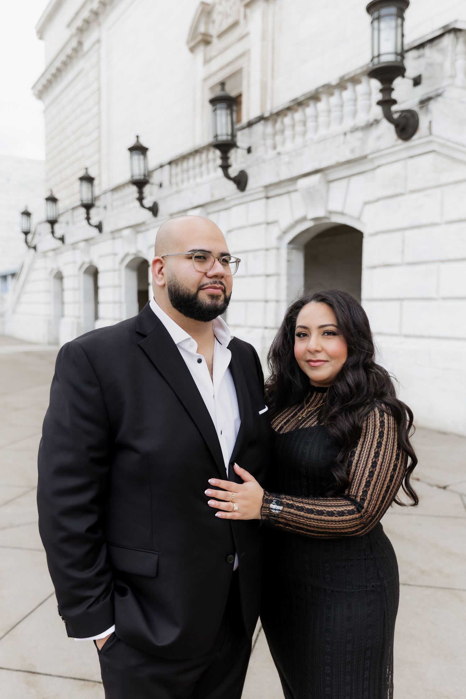 A stylishly dressed couple posing in front of a white stone building with elegant lamp fixtures.