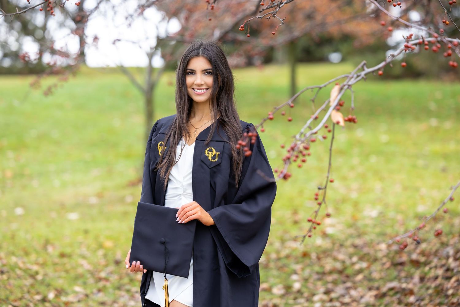 A graduate in a black academic gown stands outdoors with blooming trees in the background.