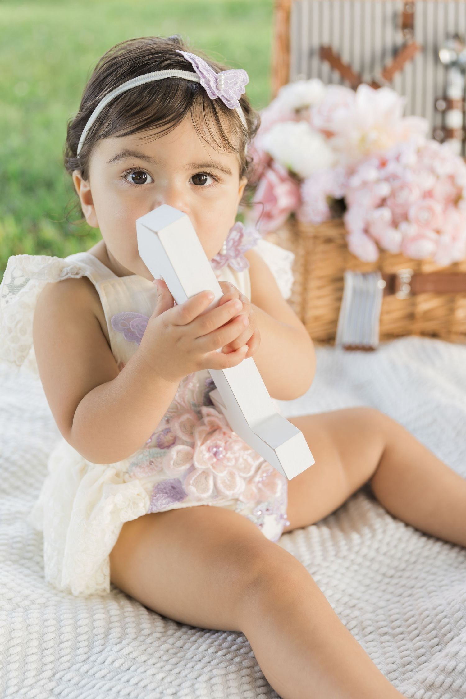 A baby in a white dress sits on a blanket with cotton candy flowers while holding a bottle at a styled picnic.