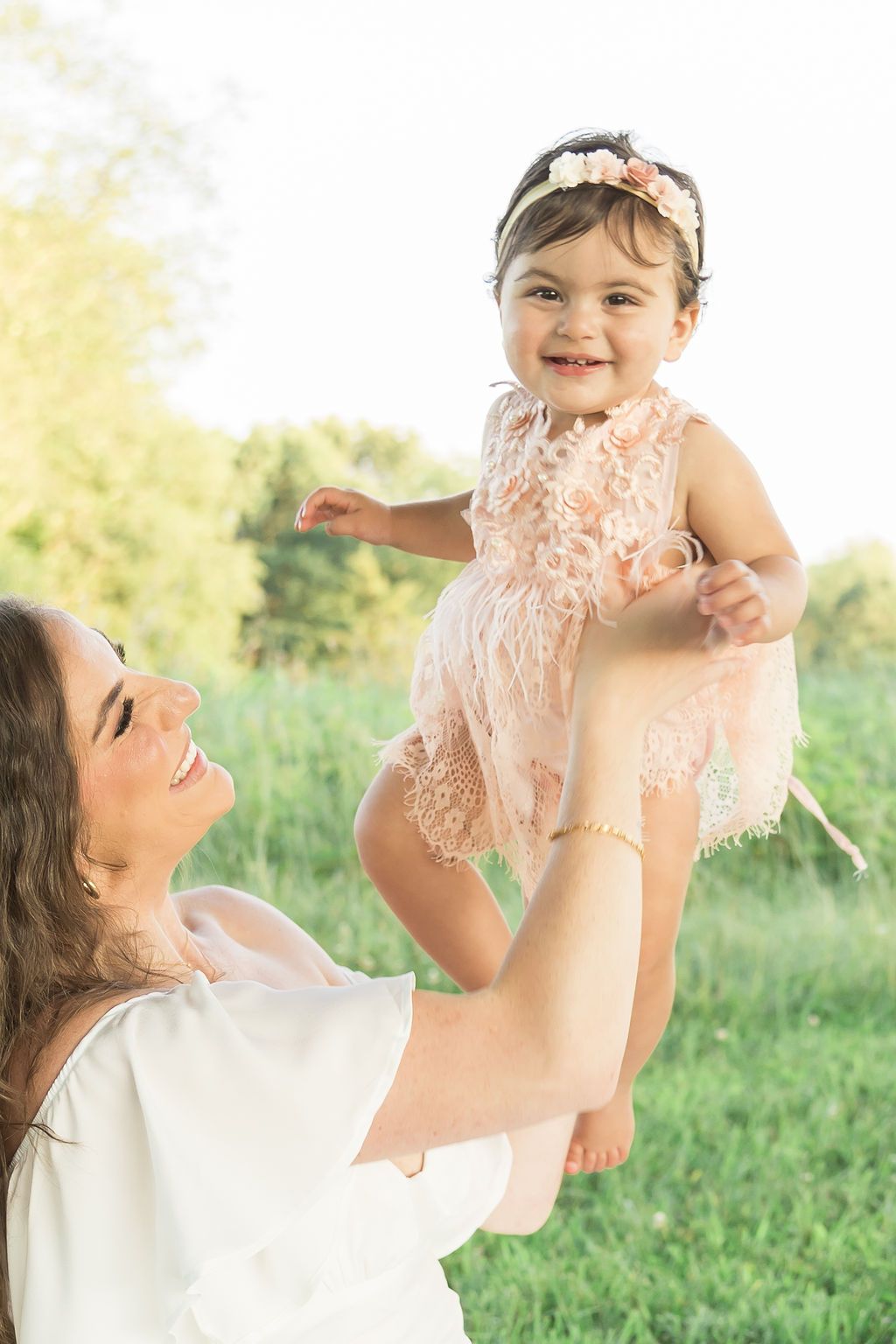 A joyful parent lifting their baby wearing a delicate peach lace dress in a sunlit grassy field.