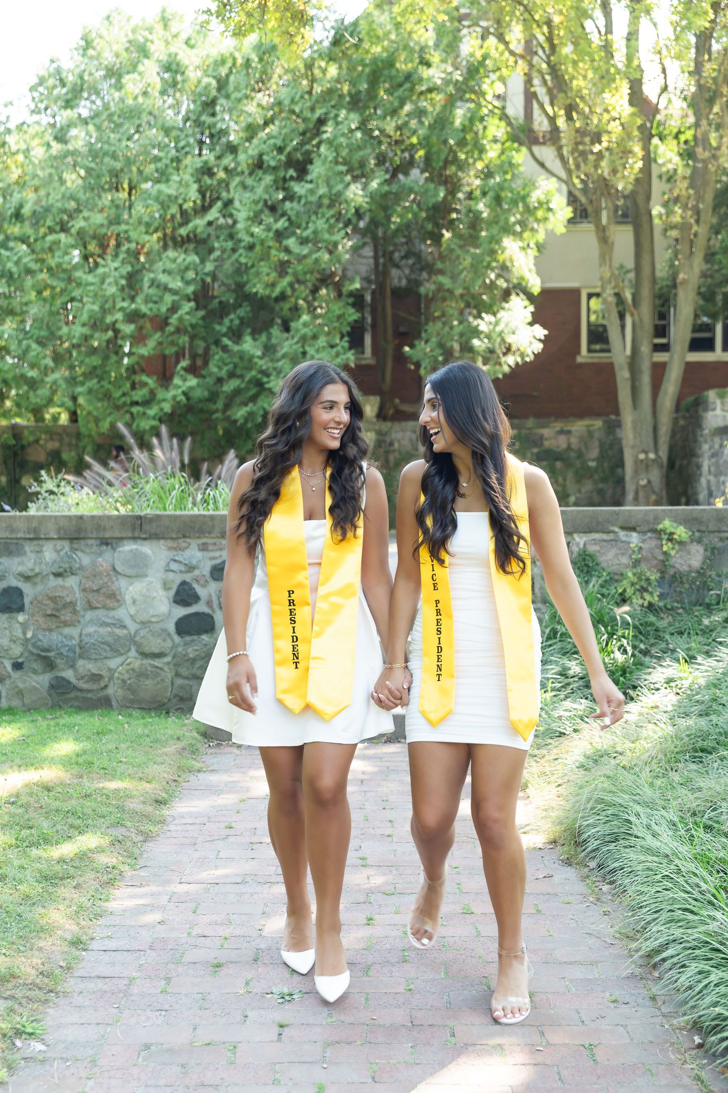 Two graduates in white dresses with yellow stoles walking together on brick path.