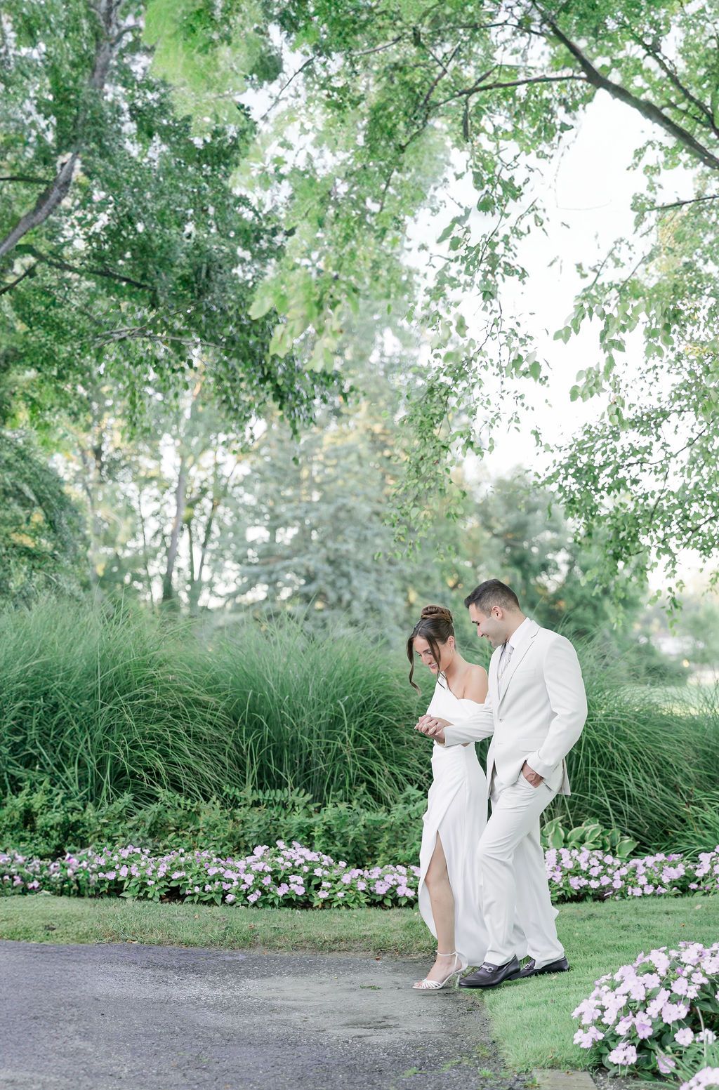 A couple in white attire walk together along a garden path bordered by pink flowers and lush greenery.